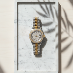 two-tone stainless steel and yellow gold Rolex Datejust luxury watch featuring a custom diamond bezel, iced out dust dial, and silver Roman numerals on a marble tray by SB Jewelers.