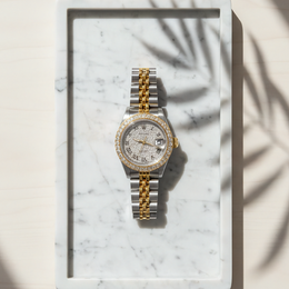 two-tone stainless steel and yellow gold Rolex Datejust luxury watch featuring a custom diamond bezel, iced out dust dial, and silver Roman numerals on a marble tray by SB Jewelers.